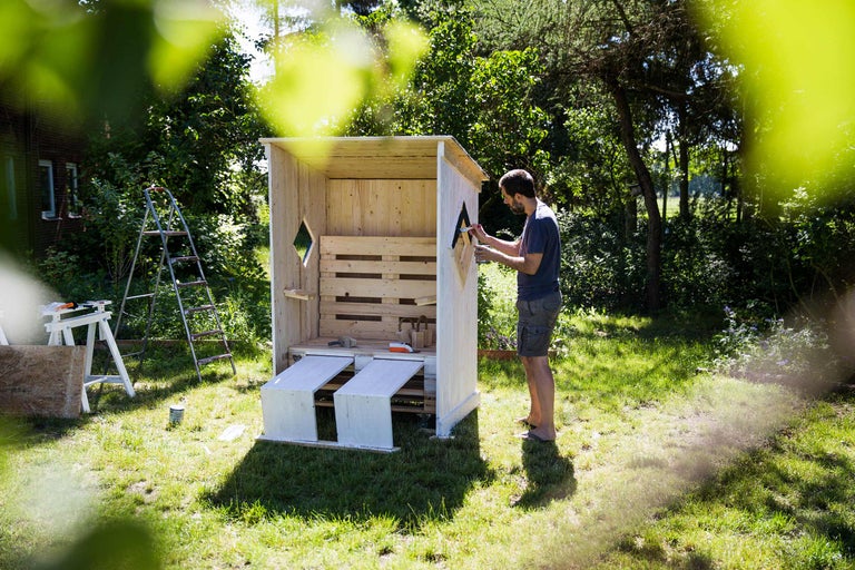 Un homme peint un abri de jardin en bois dans le jardin. Des arbres et une échelle sont visibles à l'arrière-plan.