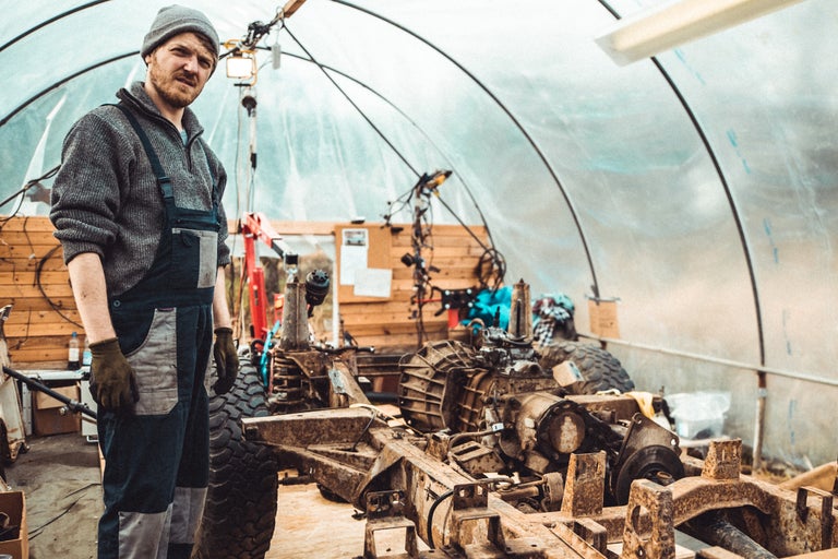 Mann steht in einer Werkstatt mit einem teilweise demontierten Geländewagen.