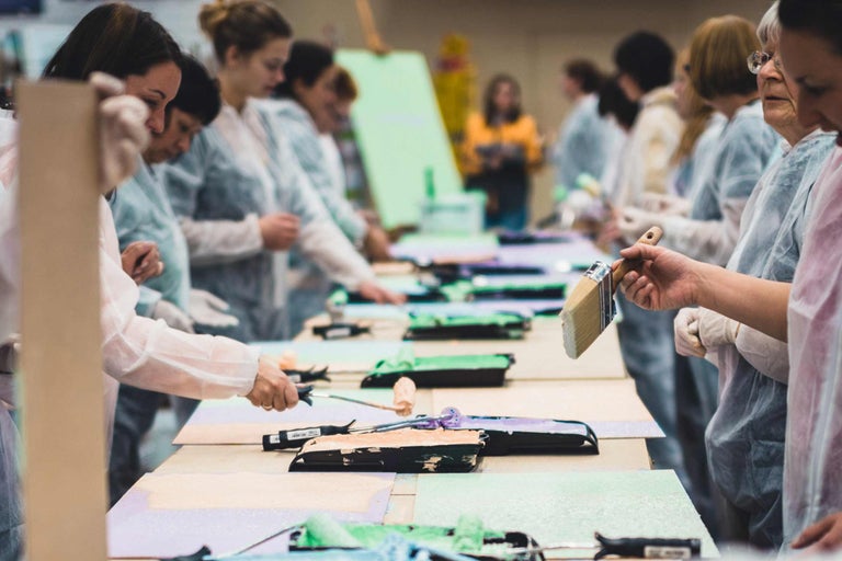 Plusieurs personnes se tiennent à des tables avec des bacs à peinture, des pinceaux et des rouleaux de peinture pour un atelier de peinture.