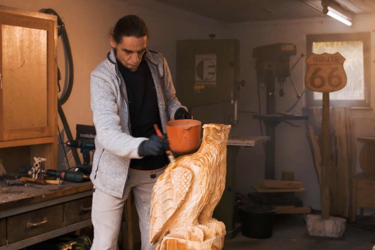 Un homme peint un hibou en bois dans un atelier. On aperçoit des outils et une perceuse en arrière-plan.