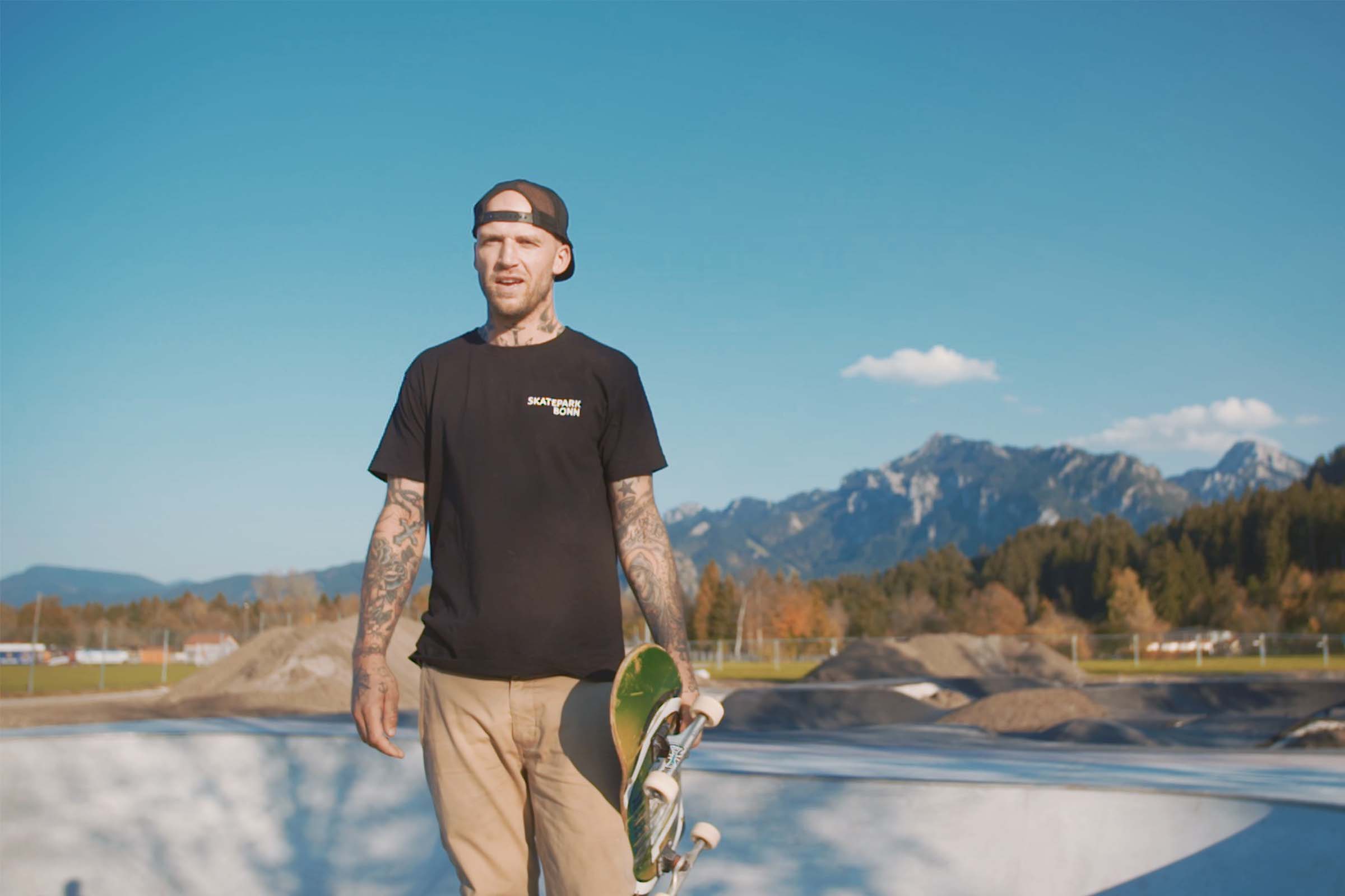 Un homme avec des tatouages tient un skateboard dans un skatepark.