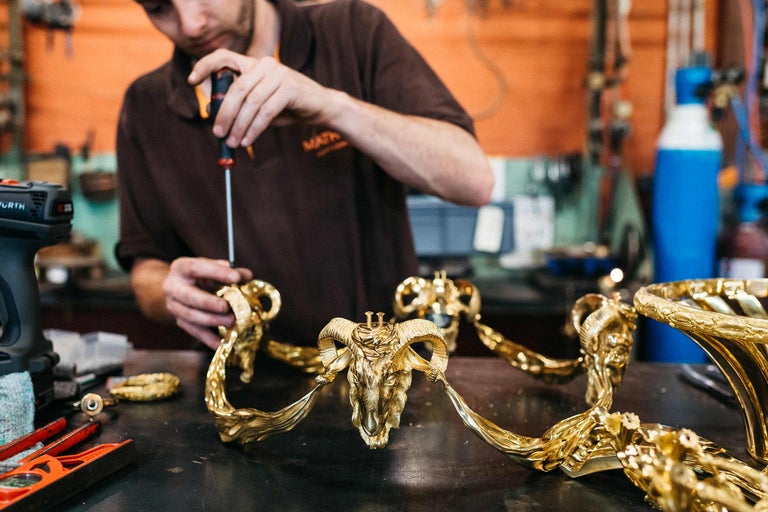 Un artisan assemble un élément décoratif avec un tournevis. L'élément représente une tête de bélier et est en métal.