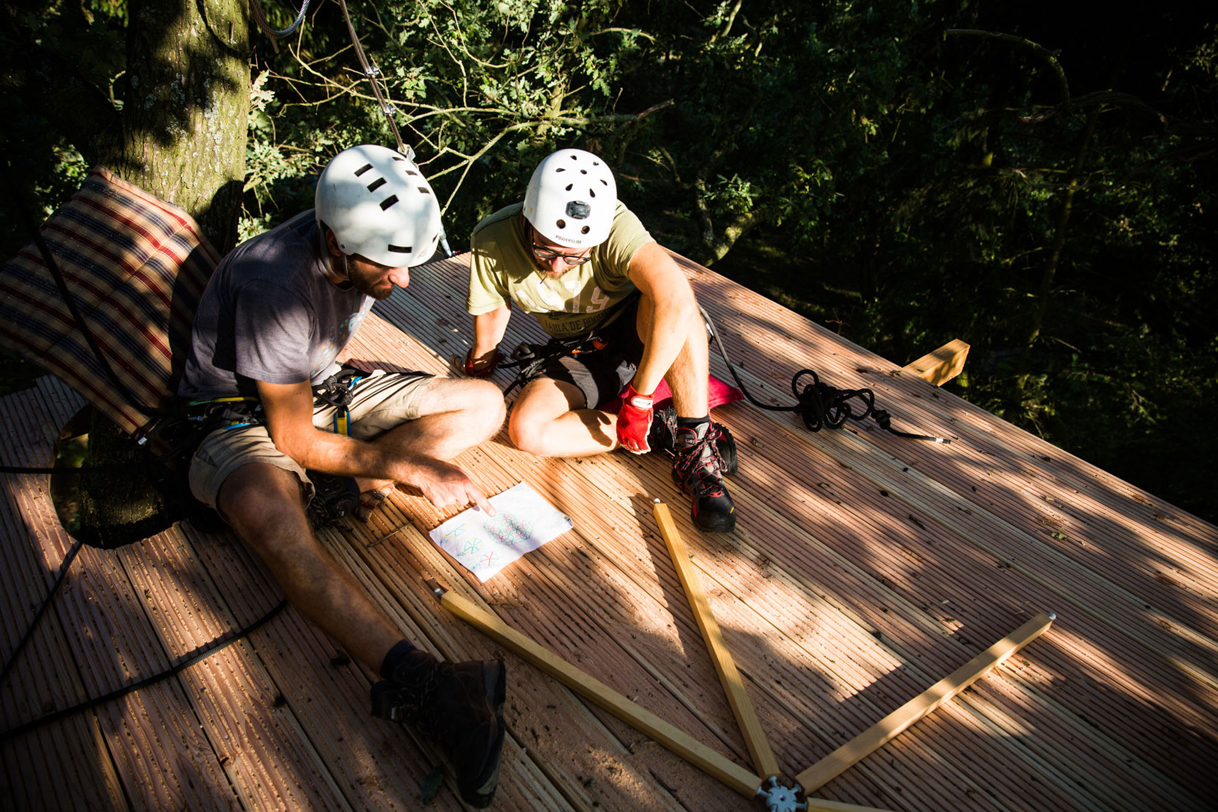 Zwei Personen mit Helmen und Sicherheitsausrüstung sitzen auf einer Holzplattform und betrachten eine Zeichnung für den Baumhausbau.