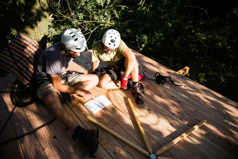 Zwei Personen mit Helmen und Sicherheitsausrüstung sitzen auf einer Holzplattform und betrachten eine Zeichnung für den Baumhausbau.