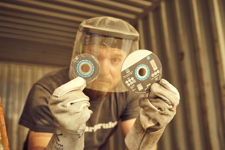 Un homme portant une protection faciale et des gants de travail tient deux disques à tronçonner.