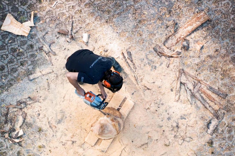 Vue de dessus d'une personne travaillant un morceau de bois avec une tronçonneuse, entourée de sciure.