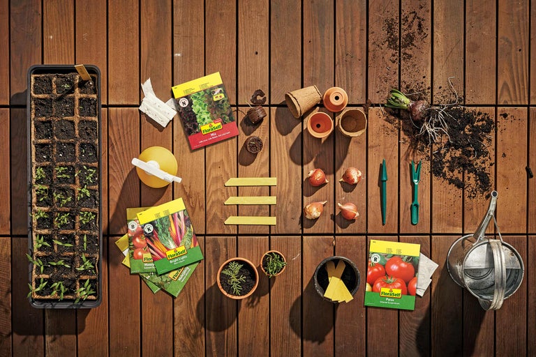 Scène de jardin avec un plateau de culture, des sachets de graines FloraSelf, des pots de fleurs et des outils de jardinage sur un plancher en bois.