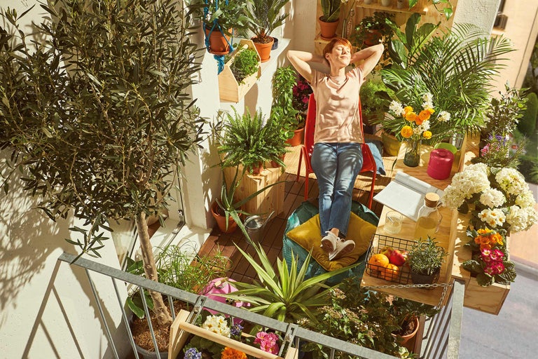 Une femme se détend sur un balcon verdoyant, entourée de plantes en pots et en bacs, avec une table avec des boissons et des fruits.