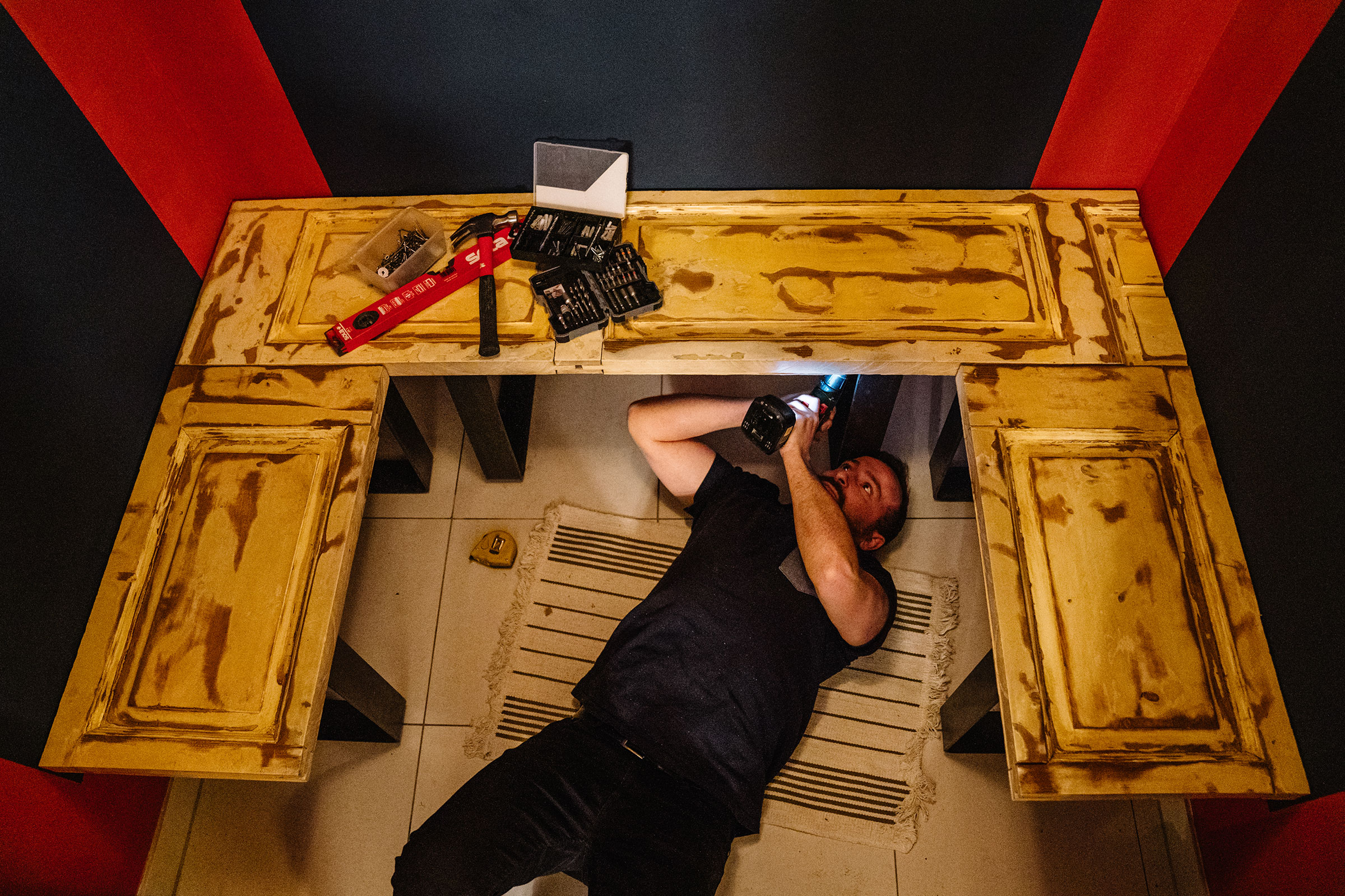 Un homme est allongé sur un tapis et monte un meuble avec un tournevis sans fil. Des outils sont à portée de main.