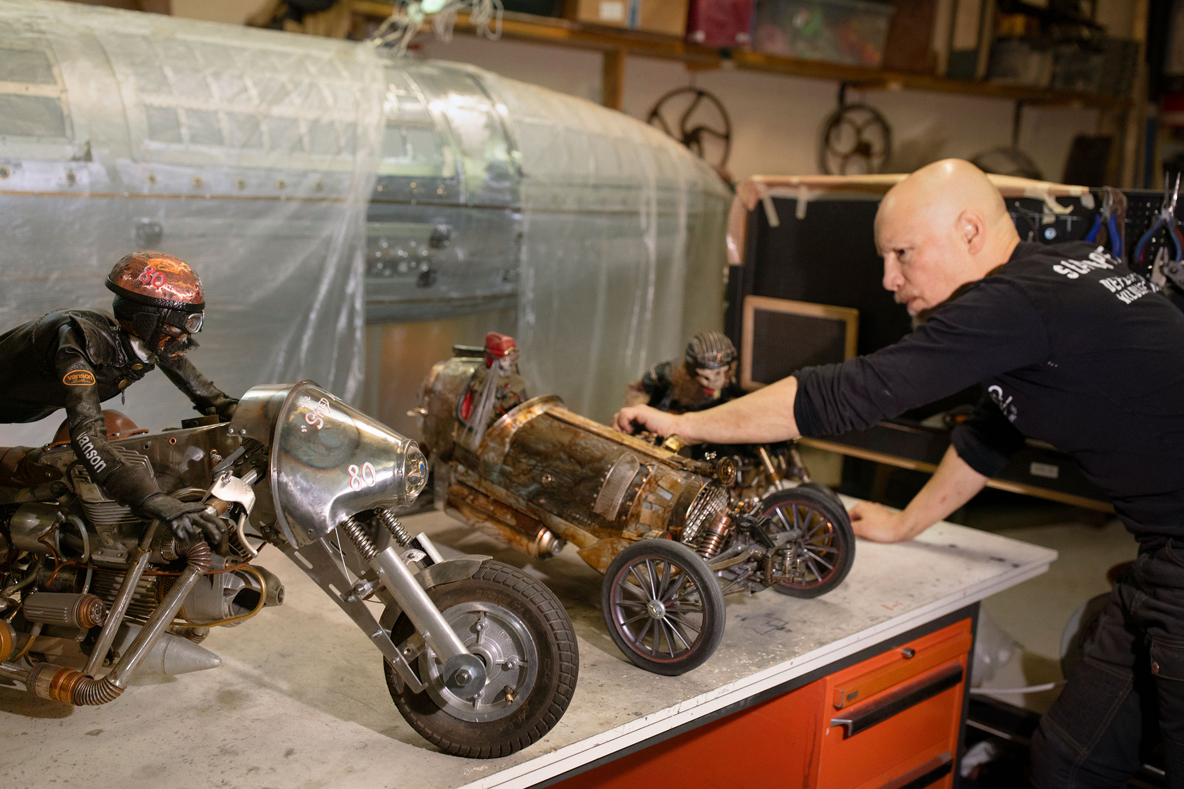 Un homme présente une moto miniature et une voiture de course miniature en métal sur un établi.