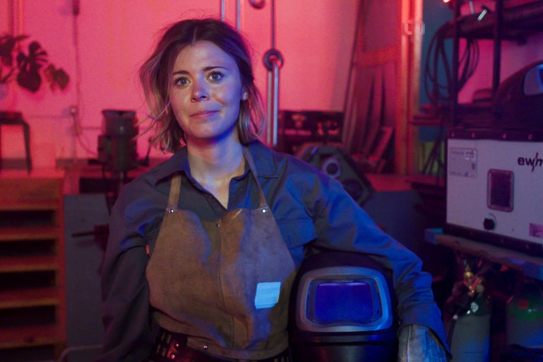 Frau mit Schweißhelm, Lederschürze und Handschuhen in einer Werkstatt