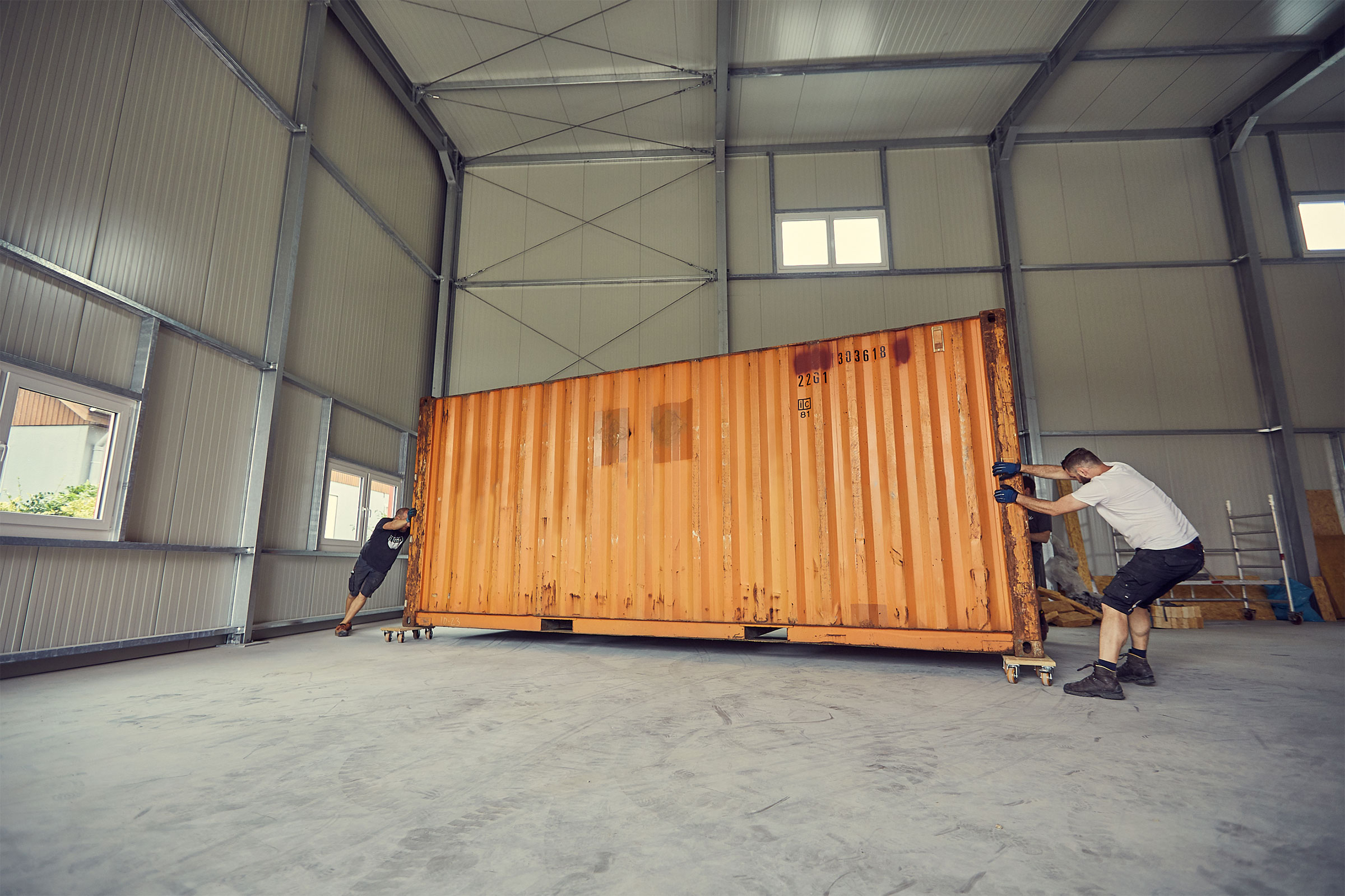 Zwei Personen bewegen einen Lagercontainer auf Rollen in einem Lagerhaus mit Metallstruktur.