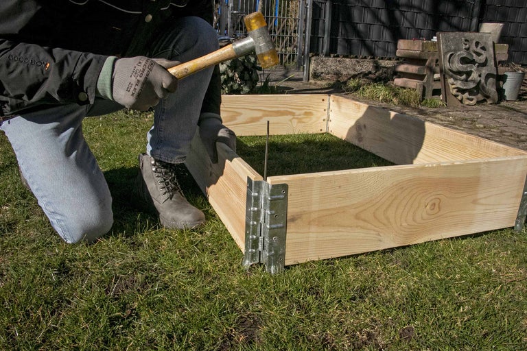 Person montiert ein Hochbeet aus Holz mit einem Hammer und Winkeln im Garten.