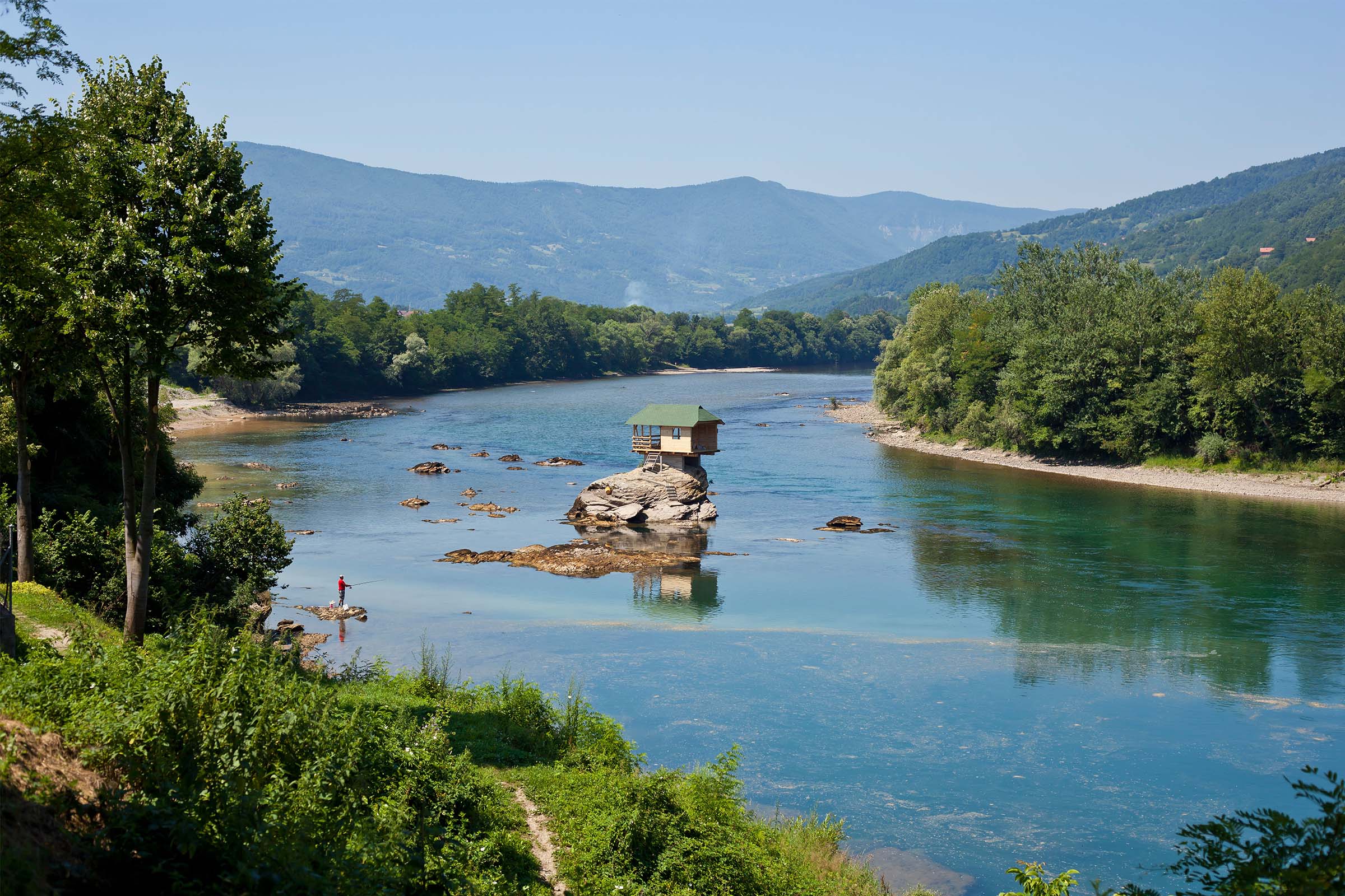 Maison sur un rocher dans la rivière avec une personne, entourée d'arbres et de montagnes