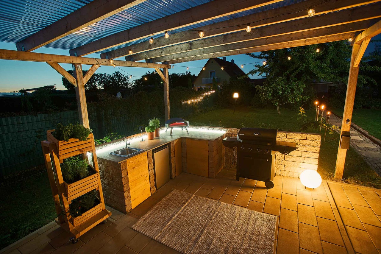 Cuisine extérieure éclairée avec barbecue, évier et étagère à plantes sous une pergola avec guirlande lumineuse