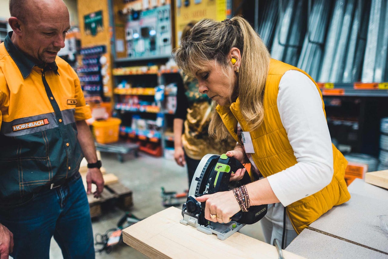 Eine Frau schneidet Holz mit einer Handkreissäge, während ein Mann im Hornbach-Outfit zusieht.