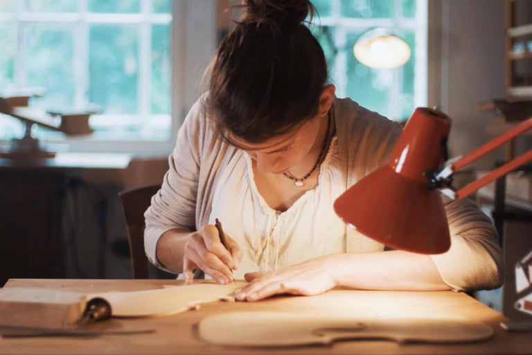 Eine Frau bearbeitet ein Holzstück mit einem Werkzeug an einem Tisch.