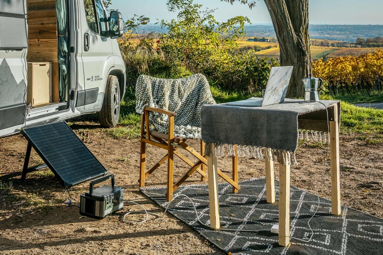 Szene im Freien mit Camper, Solaranlage, Tisch, Stuhl und Laptop in einer natürlichen Umgebung.