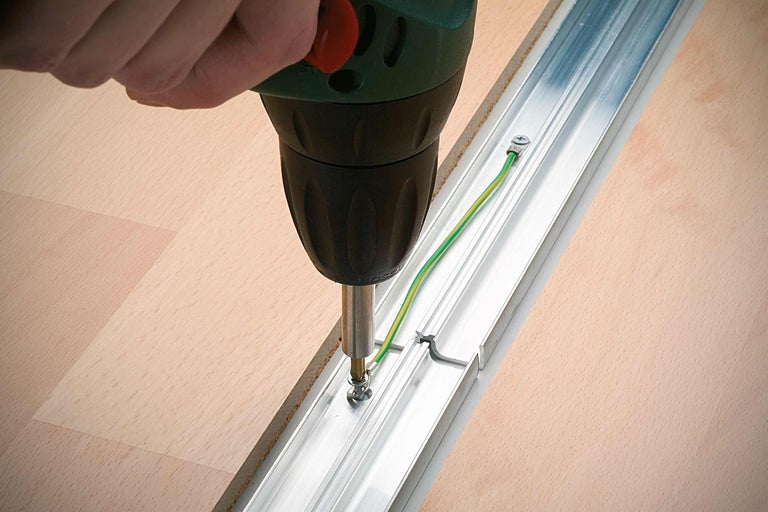 Une personne visse un rail profilé en aluminium sur un plancher en bois avec une perceuse sans fil.