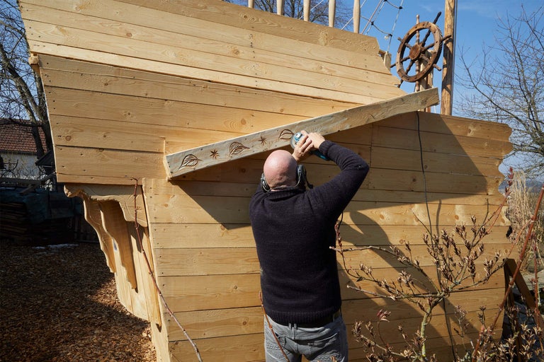 Mann bearbeitet Holz an einem Spielschiff mit einem Winkelschleifer