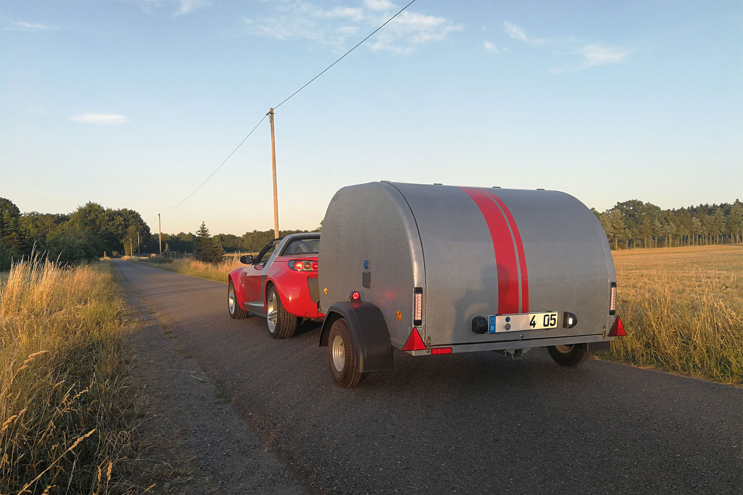 Une voiture rouge tracte une remorque grise à rayures rouges sur une route.