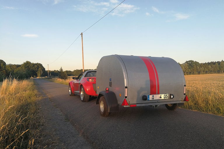Une voiture rouge tracte une remorque grise à rayures rouges sur une route.