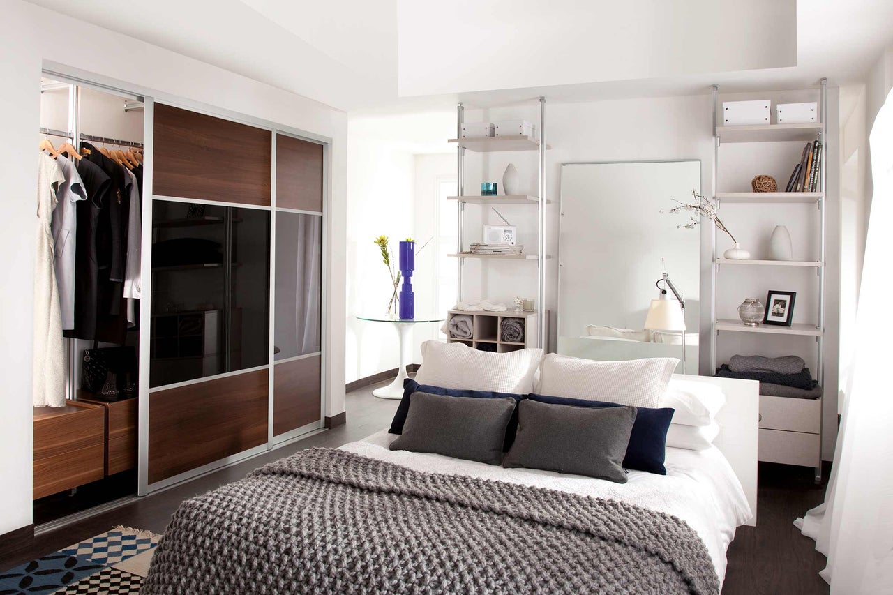 Helles Schlafzimmer mit Schiebetürenschrank, Regalen und einem Bett mit Kissen und Decke.