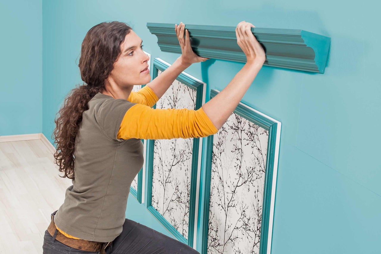 Frau montiert eine Zierleiste an der Wand über zwei gerahmten Bildern.