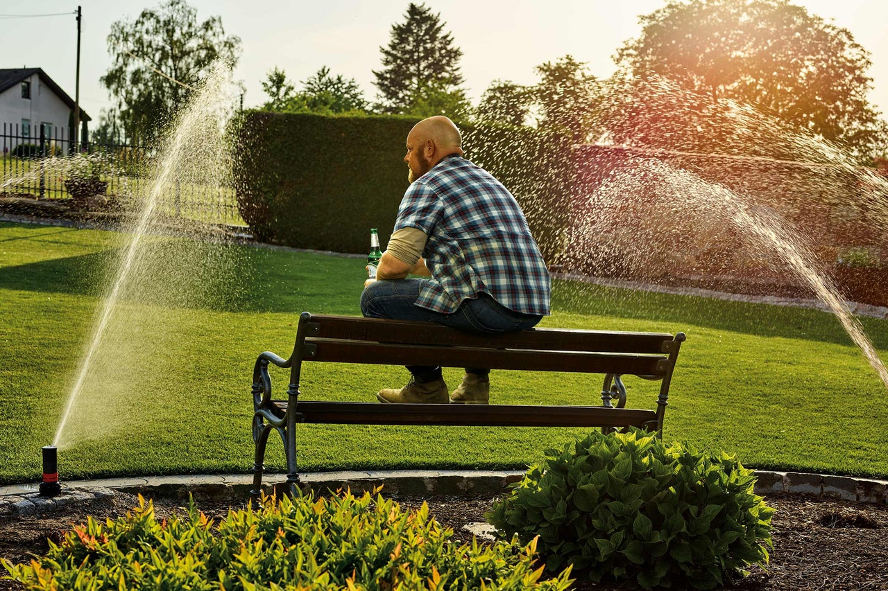 Mann sitzt auf einer Bank im Garten, während ein Rasensprenger den Rasen bewässert.