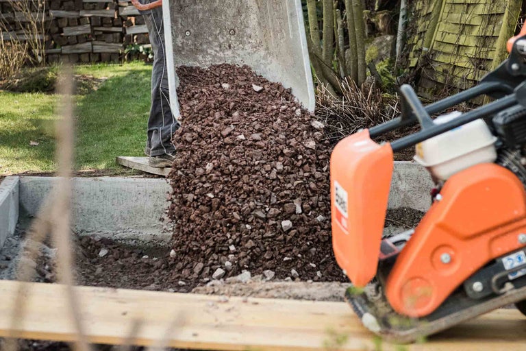 Une brouette est déversée dans une excavation, tandis qu'une plaque vibrante est prête