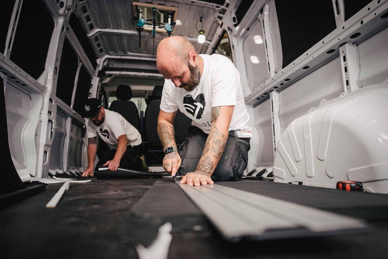 Deux artisans coupent des panneaux isolants à la taille dans l'espace intérieur d'une camionnette.