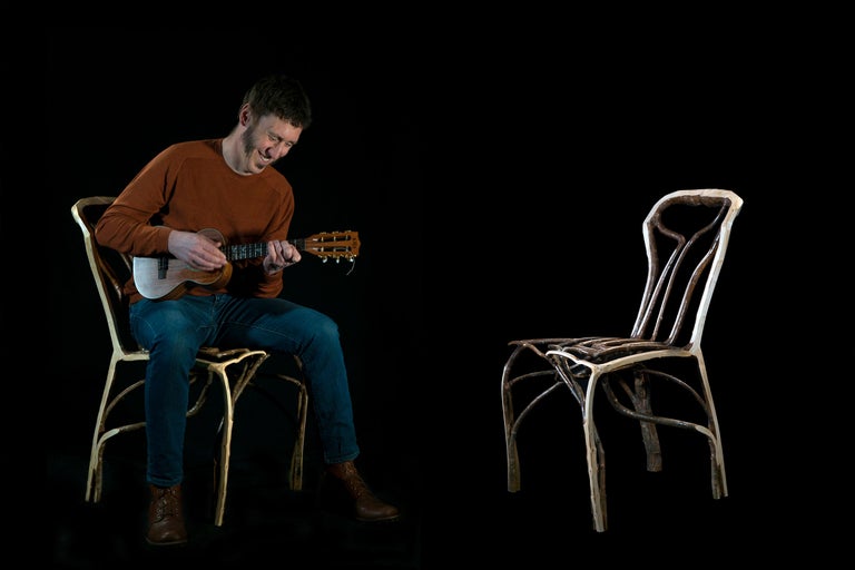 Un homme est assis sur une chaise et joue du ukulélé, une chaise vide se trouve à côté