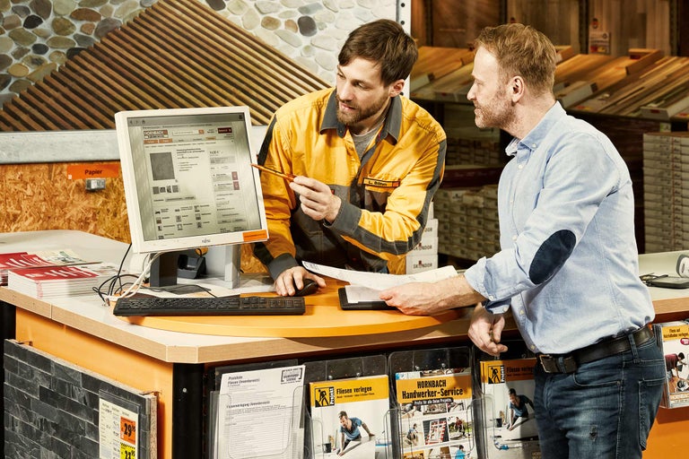 Deux collaborateurs se concertent à une table de conseil chez Hornbach, sur laquelle se trouve un ordinateur avec le site web de Hornbach.