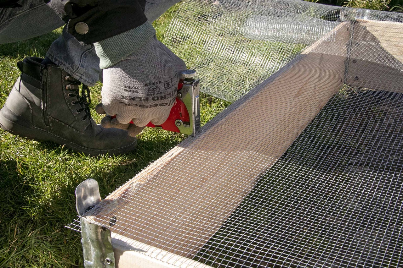Une personne fixe un grillage à l'aide d'une agrafeuse sur un cadre en bois.