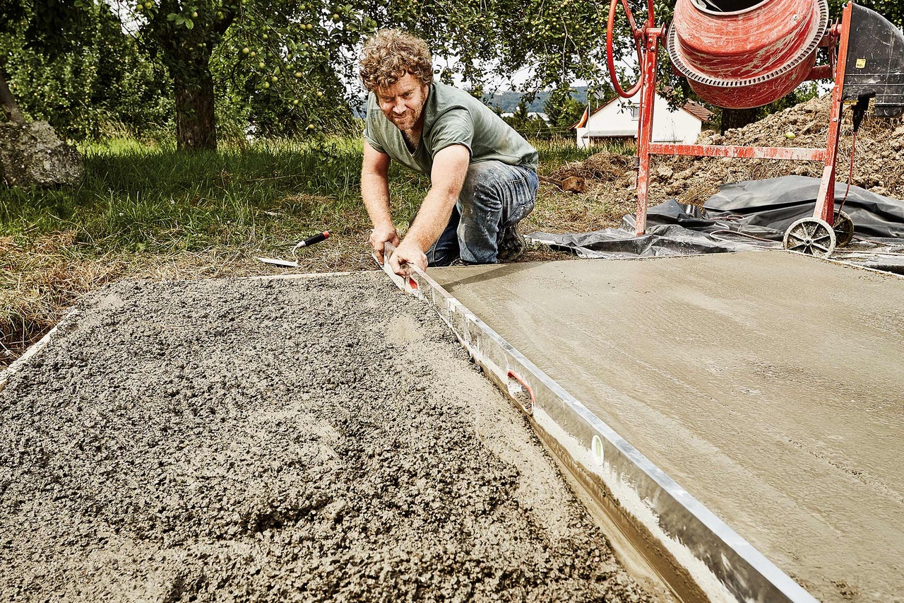 Un homme égalise du béton avec un niveau à bulle, avec une bétonnière en arrière-plan.