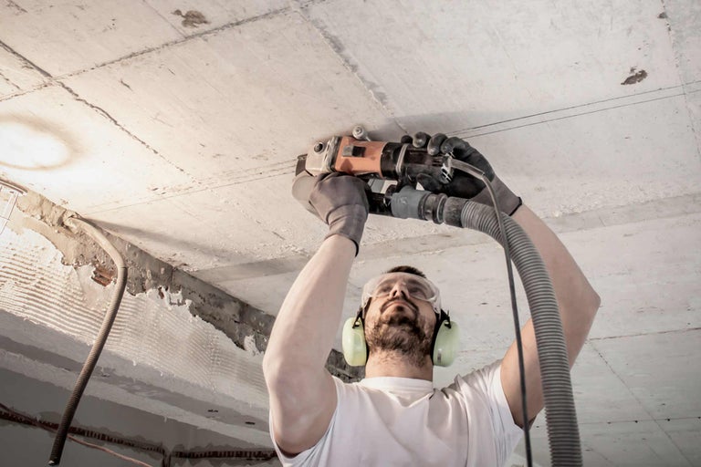 Un homme ponce un plafond en béton avec une ponceuse à béton, portant des lunettes de protection et un casque antibruit