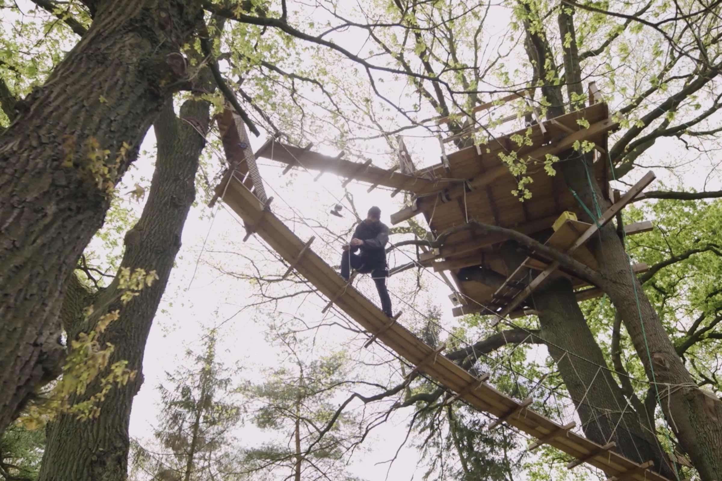 Cabane dans les arbres avec pont suspendu et une personne sur le pont.