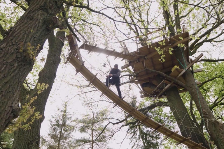 Baumhaus mit Hängebrücke und einer Person auf der Brücke.