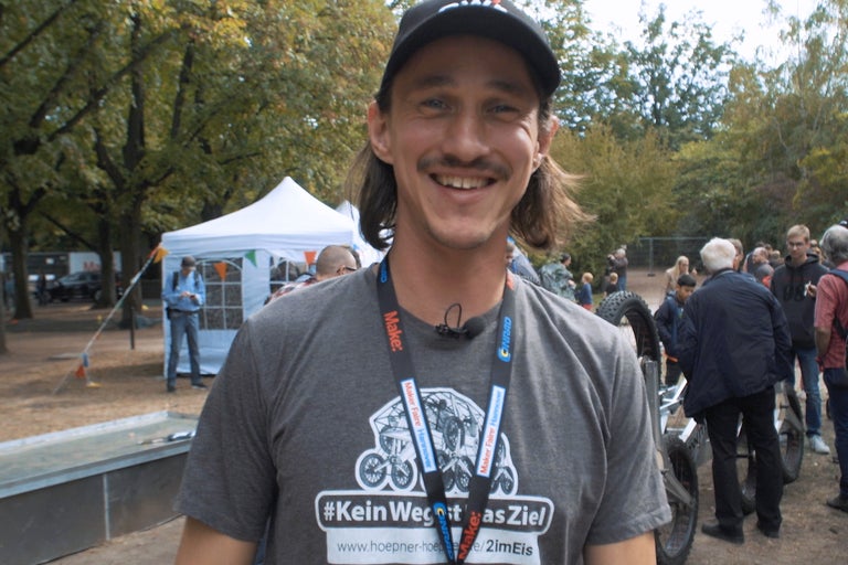 Portrait d'un homme souriant avec une moustache et une casquette, portant un t-shirt avec l'inscription '#KeinWegItAsZiel' et un lanyard de Maker Faire Hannover