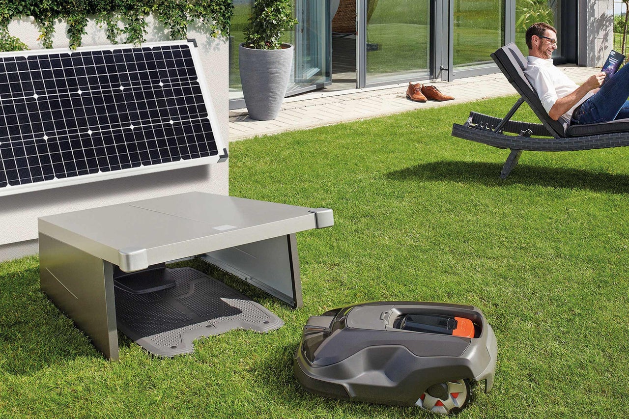 Scène avec un robot tondeuse à gazon, une station de charge avec panneau solaire et une personne se détendant sur une chaise longue dans le jardin.