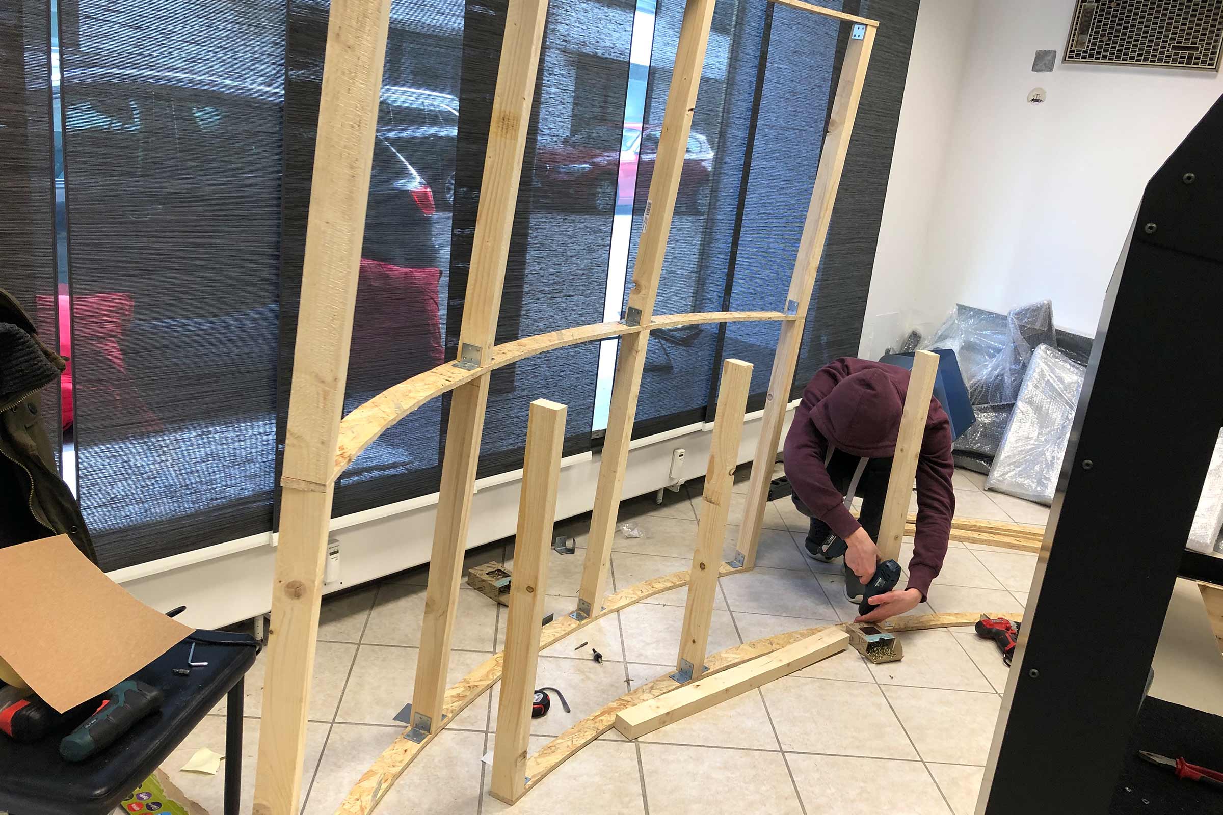 Un homme assemble une construction en bois avec une visseuse sans fil dans un intérieur.