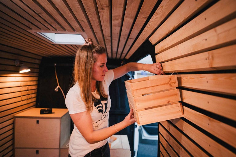 Une femme fixe une caisse en bois sur un mur en panneaux de bois dans un camping-car.