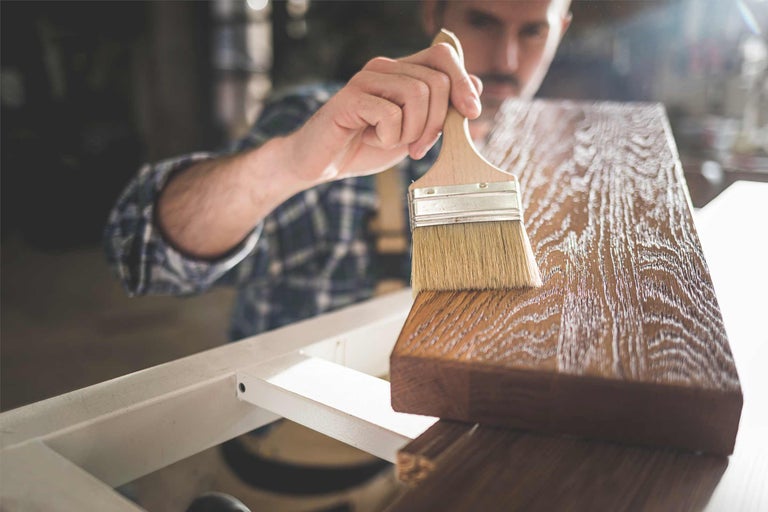 Eine Person trägt mit einem Pinsel eine Holzschutzlasur auf ein Holzbrett auf.
