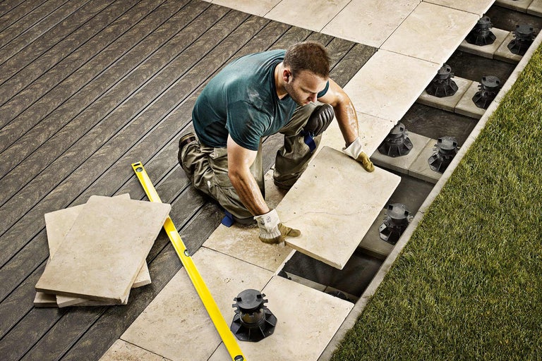 Un homme pose des dalles de pierre sur une terrasse avec des plots réglables et un niveau à bulle.