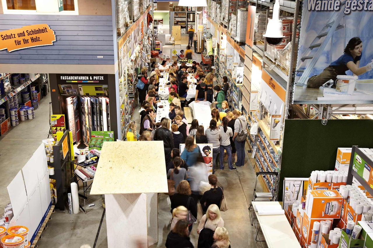 Innenaufnahme eines Hornbach Baumarktes mit Kunden, die an Tischen sitzen, umgeben von Produkten wie Farben, Putze und Tapeten.