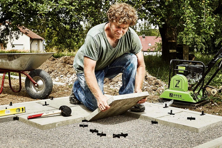 Un homme pose des dalles de trottoir avec une plaque vibrante, une brouette, un maillet en caoutchouc et un niveau à bulle dans le jardin.