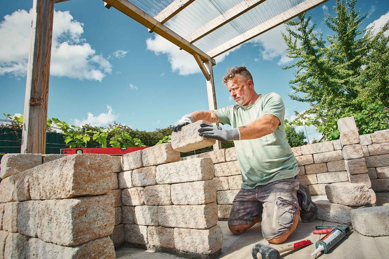 Ein Mann baut eine Gartenmauer aus Stein unter einer Holzkonstruktion mit durchsichtigem Dach.