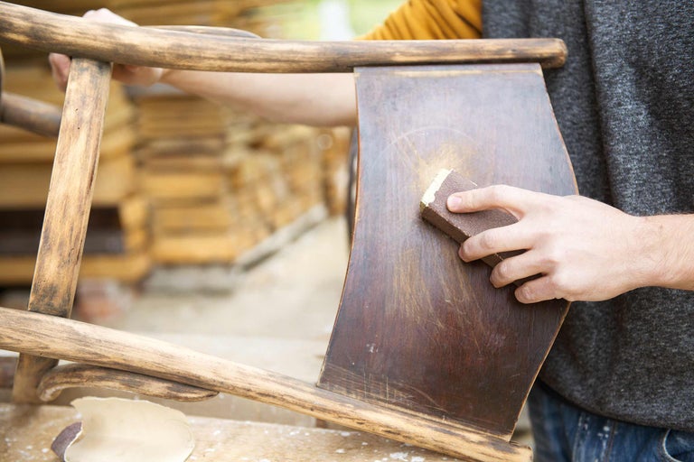 Eine Person schleift einen Holzstuhl mit Schleifpapier ab. Der Stuhl ist aus Holz und hat eine dunkle Oberfläche.