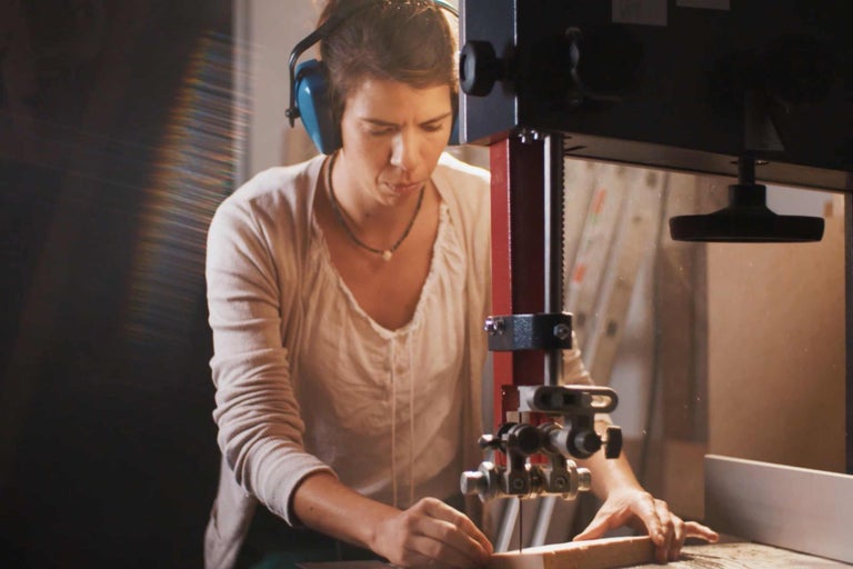 Eine Frau sägt mit einer Bandsäge ein Stück Holz, während sie einen Gehörschutz trägt.