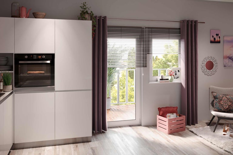 Cuisine claire avec four encastrable, fenêtre donnant sur un balcon avec des stores et des rideaux, ainsi que des décorations.